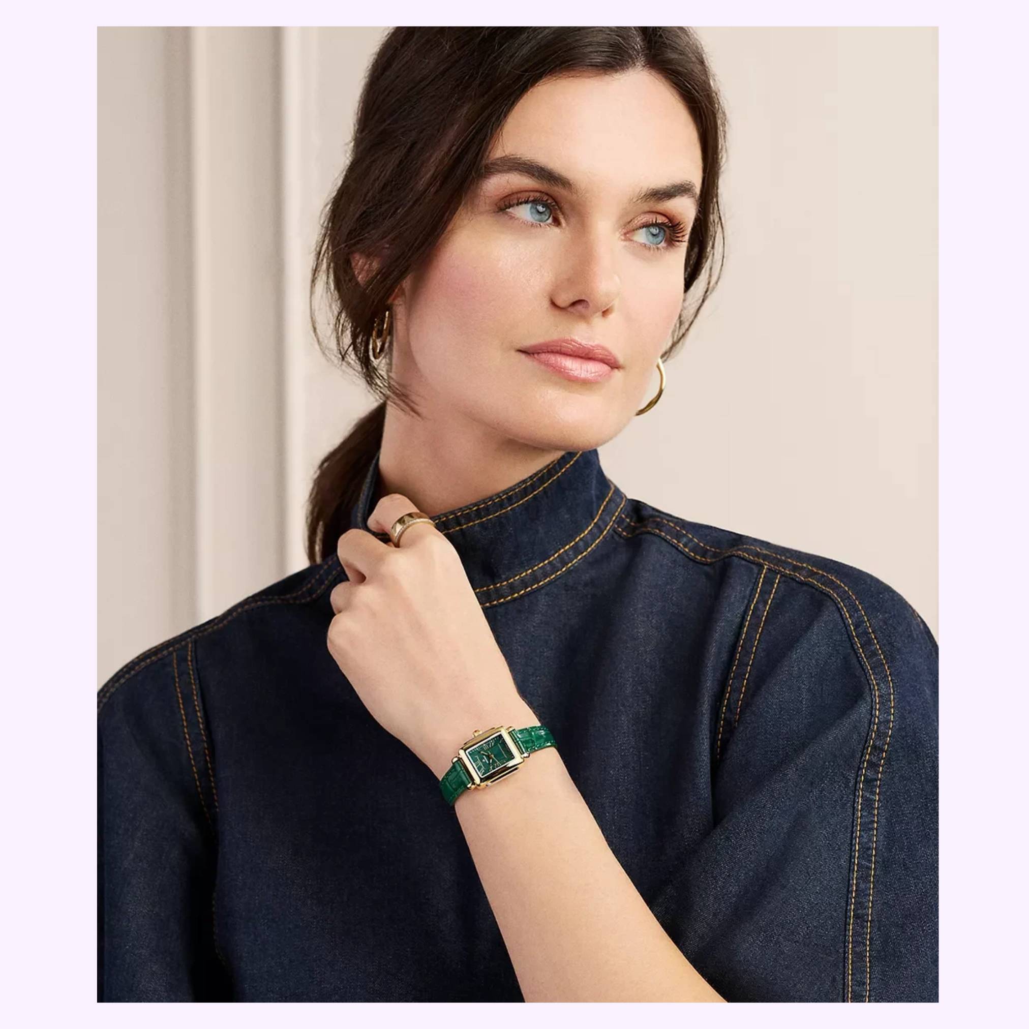 A woman in a dark denim top looks to the side, wearing a gold hoop earring, a gold ring, and the Michele Watches Deco Madison Mini MWW06D000182 green leather watch by Michele Watches on her wrist.