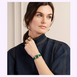 A woman in a dark denim top looks to the side, wearing a gold hoop earring, a gold ring, and the Michele Watches Deco Madison Mini MWW06D000182 green leather watch by Michele Watches on her wrist.