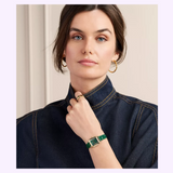 A woman with brown hair styled in a low bun wears a dark denim shirt, gold hoop earrings, a gold ring, and the Michele Watches Deco Madison Mini MWW06D000182 by Michele Watches, featuring a green leather strap and gold case. She rests her hand near her chin while looking at the camera.