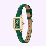 A rectangular Michele Watches Deco Madison Mini MWW06D000182 wristwatch in gold-tone, featuring a green dial with Roman numerals, a matching green leather strap, a red crown, and silver side accents. The watch is displayed against a light purple background.