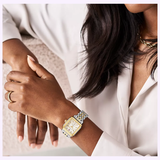 A woman with medium skin tone wears a white blouse, gold jewelry, and the Michele Watches Deco Mid Two-Tone 18K Gold-Plated Diamond Dial MWW06V000042 wristwatch with a square face, resting her arm on a light surface.