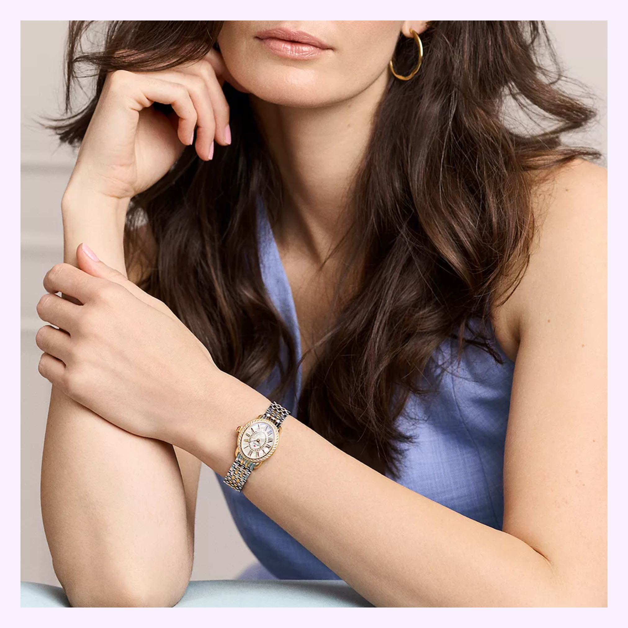 A woman with wavy brown hair, wearing a sleeveless light blue top, gold hoop earring, and the Michele Watches Serein Oval Diamond Two-Tone 18K Gold-Plated Watch (MWW21H000002), rests her hand on her chin, gazing to the side.