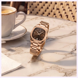 A Mido Watches Multifort TV 28 Quartz M0491103329600 in gold is displayed upright on a marble café table beside a cup of coffee and a napkin, with reflections of wicker chairs in the background.