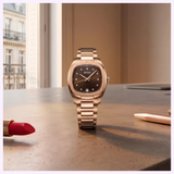 A gold Mido Multifort TV 28 Quartz M0491103329600 wristwatch by Mido Watches is displayed on a brown desk near a red lipstick, a gold pen, and a window overlooking classical buildings, exuding refined elegance.