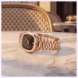 A Mido Multifort TV 28 Quartz M0491103329600 by Mido Watches, featuring a rose-gold-coloured PVD case and a sleek black dial, rests on an outdoor marble café table. In the background, woven chairs and a cup with saucer evoke a relaxed, elegant atmosphere.