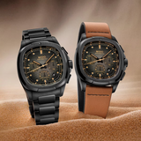 Two Mido Watches Multifort TV Chronograph (M0495273308100) sit on sand—one with a black metal bracelet, the other with a brown leather strap. Both have black dials with gold accents, black cases, and offer a 60-hour power reserve.