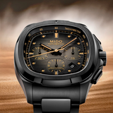 The Mido Watches Multifort TV Chronograph (M0495273308100), a black Swiss automatic watch with gold accents, chronograph, tachymeter scale, date window, and 60-hour power reserve, is shown against a blurred sandy desert backdrop.