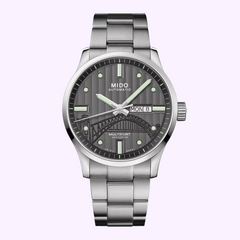 The Mido Watches Multifort IBA Limited Edition M0054301106181 features a silver case, gray patterned dial with green accents, day and date display, and a stainless steel bracelet—photographed on light purple for collectors.