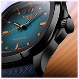 Close-up of the Mido Watches Multifort TV Big Date M0495263704100, showcasing a blue textured dial with orange markers and hands, black case, and blue fabric strap. Background is softly blurred in warm tones.