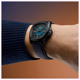 A wrist with the Mido Watches Multifort TV Big Date M0495263704100, featuring orange accents, is shown against a brown-purple gradient background. The wearer sports a navy blue sweater.