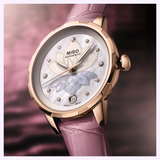The Mido Watches Rainflower Night M0432073610600, featuring automatic movement, a pink leather strap, mother-of-pearl dial with diamond markers, floral motif, and date at 6 o'clock, is displayed against a blurred pink background.