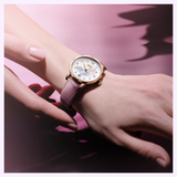 A close-up of a hand with light pink manicured nails wears the Mido Watches Rainflower Night M0432073610600, an elegant Swiss automatic women’s watch with gold accents, white dial, and light purple leather strap against a soft pink background.