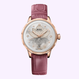 The Mido Watches Rainflower Night M0432073610600 is a Swiss women's watch featuring rose gold, an automatic movement, purple leather strap, mother-of-pearl dial with diamond markers, and date at 6 o’clock—all set against a light pink background.