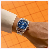 A square-cased silver Mido Watches Multifort TV Big Date Chronometer M0495671104100 wristwatch with a blue textured dial and a prominent date display worn on a wrist.