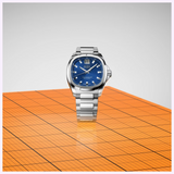 A stainless steel Mido Watches Multifort TV Big Date Chronometer M0495671104100 watch with a blue dial, square case, and a large date display at the twelve o’clock position, resting on an orange grid surface.