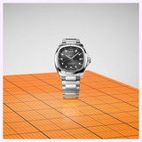 Mido Watches Multifort TV Big Date Chronometer M0495671105100 stainless steel watch with a square-edged black dial, large date display, and link bracelet, standing on an orange grid surface.