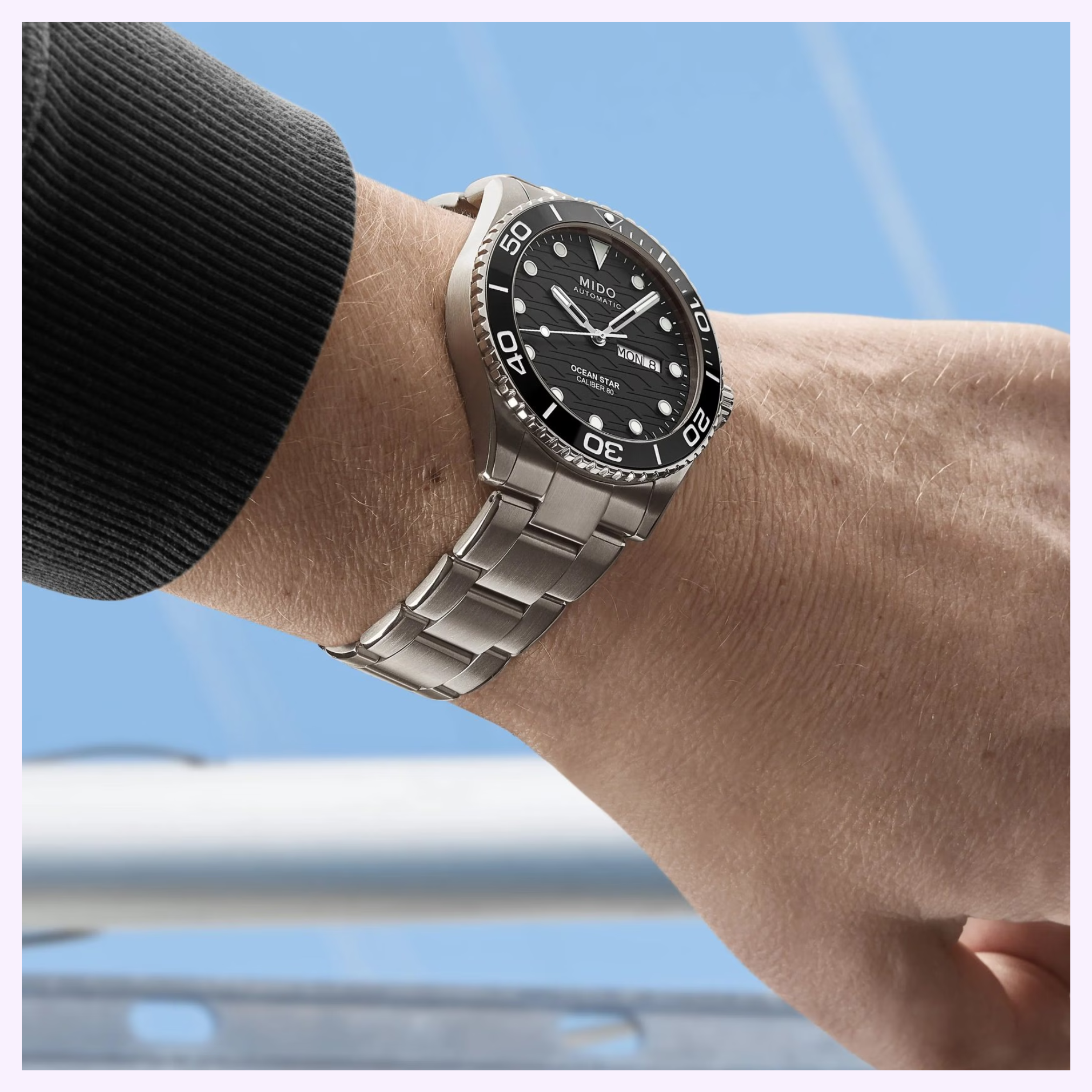 A close-up of a person’s left wrist wearing the Mido Watches Ocean Star 200C Titanium M0424304405100 with a black bezel and dial, highlighting its Swiss automatic movement against a blurred blue sky background.