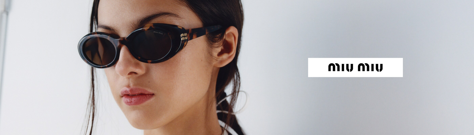 Close-up of a woman wearing dark oval sunglasses, with straight dark hair and a neutral expression, next to the Miu Miu logo on a white background.