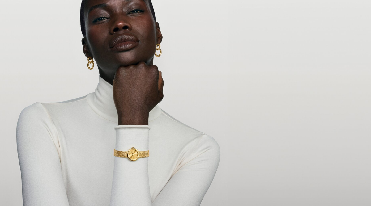A woman wearing a white turtleneck poses confidently, displaying gold jewelry including hoop earrings and a wristwatch with a round face and intricate band. The background is a smooth gradient from white to light gray.