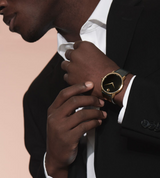 A man in a black suit adjusts his collar, wearing a Movado 0607271 MUSEUM CLASSIC Unisex Watch by Movado Watches on his left wrist against a neutral background.
