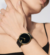 A person wears the Movado 0607271 MUSEUM CLASSIC unisex watch by Movado Watches, featuring a black and gold case with a black leather strap, paired elegantly with their black thin-strapped top.
