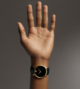 A raised hand with fingers spread, wearing a Movado Watches 0607271 MUSEUM CLASSIC unisex watch with a black leather band and round black-and-gold face, stands out against a plain gray background.