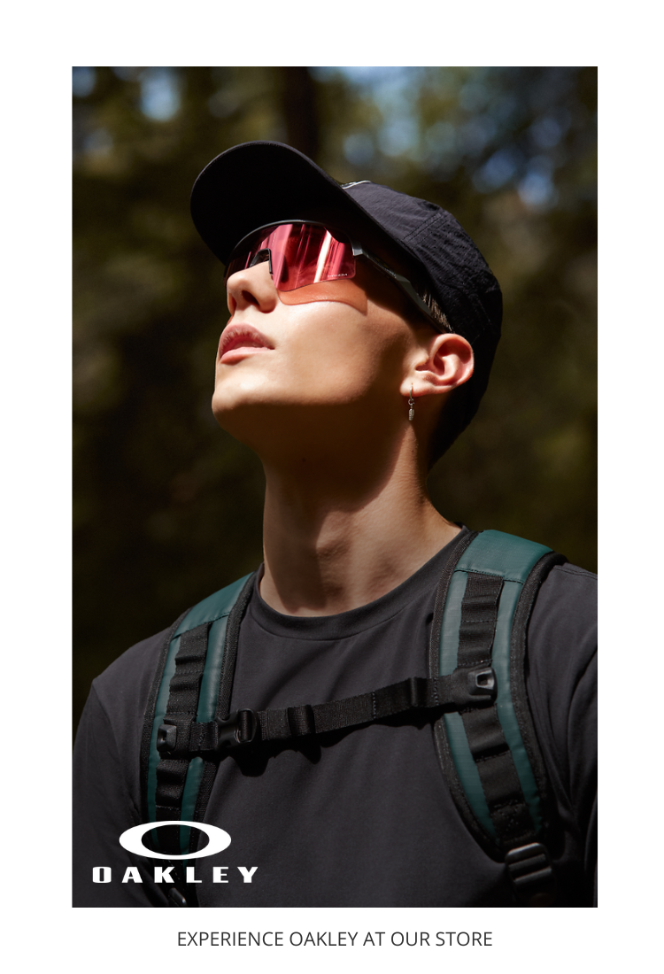 A person wearing a black cap, red-tinted Oakley sunglasses, and a dark backpack looks upwards outdoors. Oakley logo and text EXPERIENCE OAKLEY AT OUR STORE appear at the bottom.
