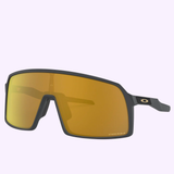 Oakley OO9406 05 37 Sutro Unisex Sunglasses with a black angular frame and large amber lens, accented by a small Oakley logo on the temple, are shown against a light purple background.