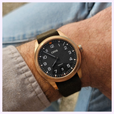 An Oris Watches ProPilot Bronze wristwatch with a bronze case, black dial, and dark green strap worn on a wrist.