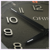 Close-up of an Oris Watches ProPilot Date dial featuring raised silver numerals, black hands with white accents, and a textured dark gray surface.