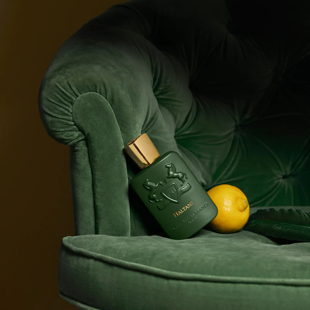 A dark green perfume bottle with a gold cap sits on a plush green velvet chair next to a yellow lemon and a dark green feather.