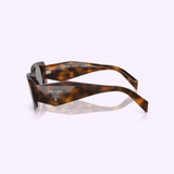The PRADA Sunglasses 0PR 17WSF 20D50Q51 by Prada Sunglasses are shown resting on their side, highlighting the thick, curved arms and the Prada Milano logo printed in white on both the inside and outside of the left arm. Set against a light background, these women’s rectangular sunglasses feature a captivating Juniper Tortoise frame that blends classic elegance with contemporary sophistication.