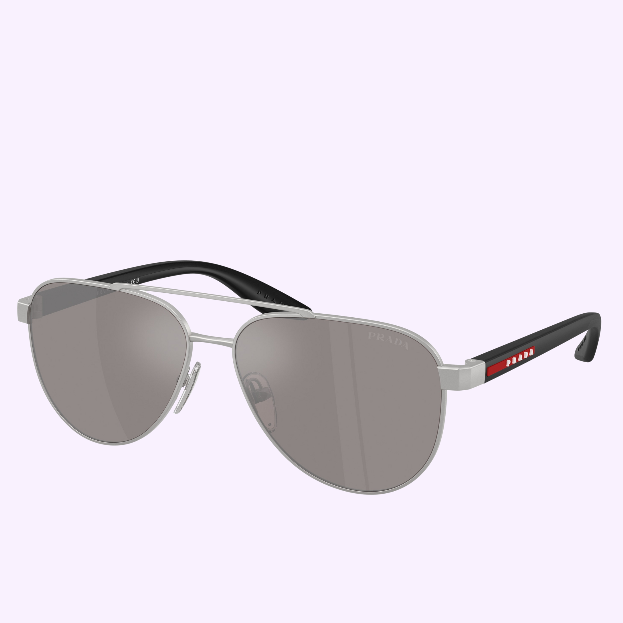 Prada Sunglasses 0PS A52S 18X80I58 feature an aviator style with silver metal frames, gray tinted lenses, black arms, and a red and white Prada logo on the right arm. Shown against a light pastel purple background.