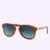 The Persol PO0714SM 96/S3 54 Unisex Sunglasses by Persol Sunglasses feature a brown tortoiseshell frame, round blue-tinted lenses, and silver hinge accents, inspired by classic Steve McQueen styles, and are showcased against a light purple background.