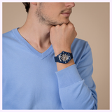 Man wearing a blue v-neck sweater with a Rado Watches Captain Cook High-Tech Ceramic Skeleton R32153209 on his wrist, mesh strap visible, as he raises his hand to his chin in a thoughtful pose.