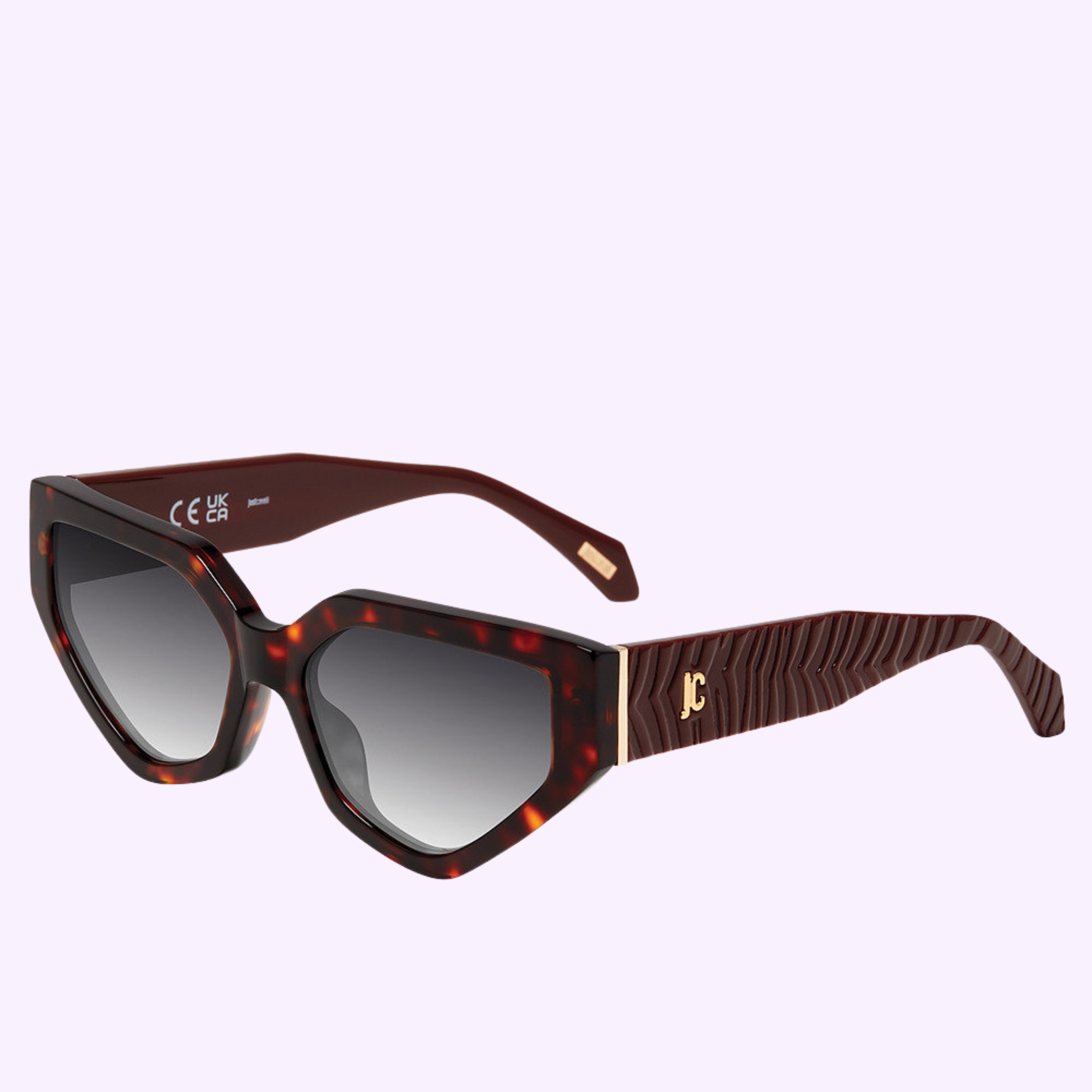 The Roberto Cavalli Sunglasses SJC196 560909 feature an angular cat-eye shape, tortoiseshell frame, dark gradient lenses, and wide textured arms with a gold JC logo. Displayed on a plain light purple background.