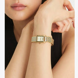 A person wearing a sleeveless black top showcases the Tory Burch Watches THE ELEANOR TBW1066 (UPC 796483611962) from Tory Burch Watches, featuring a gold rectangular face and a linked band, as they rest their chin on their crossed arms—a stylish example of a women’s stainless steel watch.