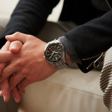 A person in a black jacket and plaid pants sits with hands clasped, wearing the Seiko Luxe Watches PRESAGE SHARP-EDGED SERIES GMT SPB221—an automatic GMT timepiece—on their left wrist.