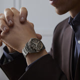A person in a dark suit sits with hands clasped, highlighting the Seiko Luxe Watches PRESAGE SHARP-EDGED SERIES GMT SPB225 on their wrist, showcasing its black dial and silver details with refined Seiko elegance.