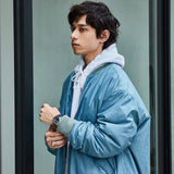 A young person with short, wavy dark hair wears a light gray hoodie under a shiny blue bomber jacket, glancing sideways and adjusting their cuff, sporting the Seiko Watches 5 Sports SKX series SRPH31 on their wrist.