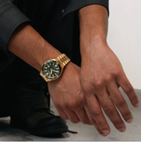 A person in dark clothing and black boots rests their hands on their knee, wearing a Citizen Watches TSUYOSA Shore NJ0232-53X by Citizen Watches—a yellow gold-tone stainless steel watch with a green face—against a plain, light-colored background.