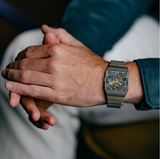 A close-up of hands resting together, wearing the Rado Watches Anatom Automatic Skeleton R10206109 with visible gears and metal band. The person is dressed in a teal long-sleeve shirt and light-colored pants.