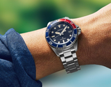 A close-up shows a wrist wearing the Seiko Watches Prospex Sea SNE595 by Seiko Luxe Watches, featuring a blue and red bezel, blue dial, and silver bracelet. The wearer is in a blue shirt against a blurred green backdrop.