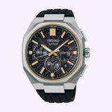 The Seiko Luxe Watches Astron HAB003 from the King Watches line features a black dial with gold accents, three subdials, a silver hexagonal titanium case, and a black rubber strap, displayed on a light purple background.