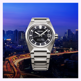 A Seiko King Watches VANAC HKF003 by Seiko Luxe Watches, with a silver case and black face, is displayed against a cityscape at dusk. Illuminated skyscrapers and winding highways with glowing car lights highlight the watch—a striking tribute to the elegance and precision of Japanese watchmaking.