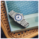 A Seiko Luxe Watches King VANAC HKF004 Limited Edition wristwatch with a blue and white face rests on a closed book with a blue-green cover, atop a woven rattan surface.