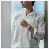 A man in a white button-up shirt stands by a window, gazing outside. He has short hair and is wearing a Seiko King Watches VANAC HKF004 Limited Edition from Seiko Luxe Watches on his left wrist. The watch’s automatic movement gleams subtly in the soft daylight that fills the space.