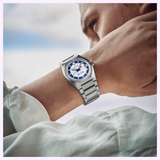 A person wearing a silver wristwatch with a blue and white face—the Seiko King Watches VANAC HKF004 Limited Edition by Seiko Luxe Watches—looks thoughtfully into the distance, their arm raised close to their face.