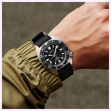 A person wearing the Seiko Luxe Watches PROSPEX AUTOMATIC DIVER'S WATCH SPB317, featuring a black strap and black dial, is dressed in an olive-green jacket. The watch, equipped with the precise Seiko 6R35 movement, displays the time as 10:13:36. The background is blurred.