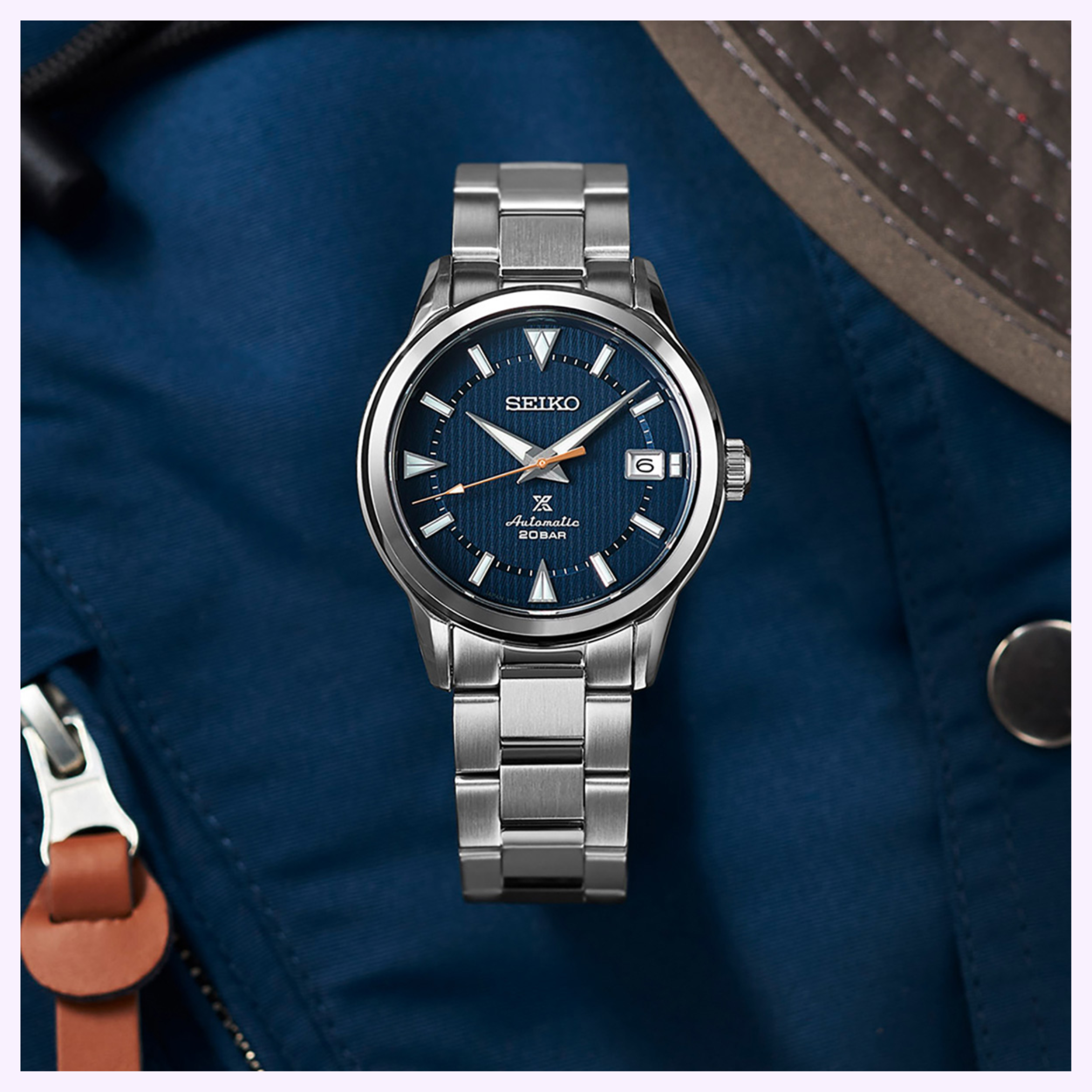 A Seiko Luxe Watches PROSPEX SPB249, featuring a blue textured dial and automatic movement, sits on a blue fabric surface next to a brown strap and part of a gray hat.